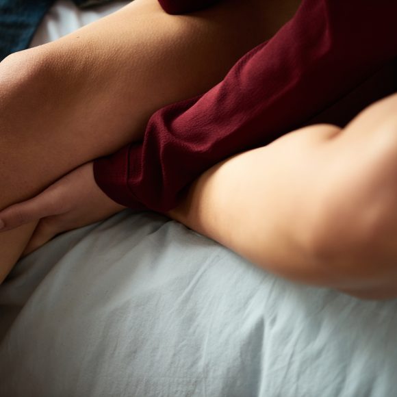 Close-up of unrecognizable woman in red blouse holding hand under her knee while sitting on bed.