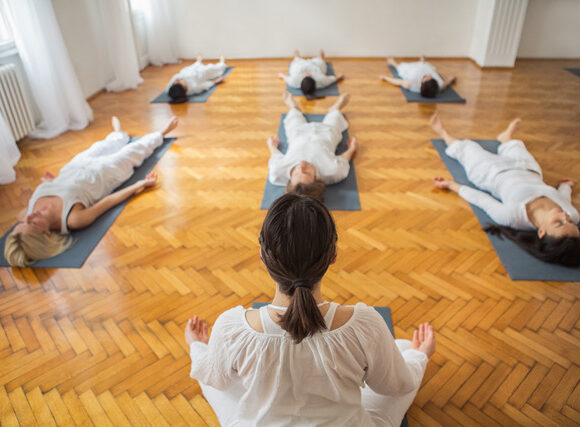 A person is leading a yoga class with participants lying on their backs on mats in a room with wooden floors, practicing relaxation or meditation.